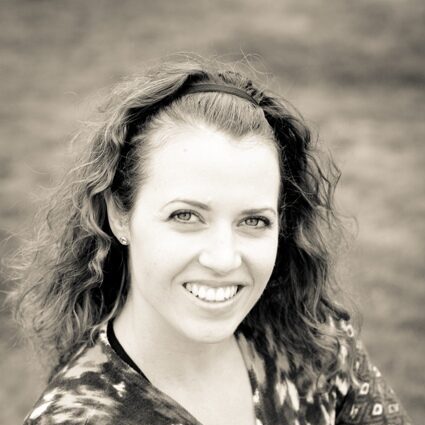 A black and white photo of a woman with long wavy hair and light skin. she is smiling at the camera and is wearing a thin hairband and a patterned v-neck top