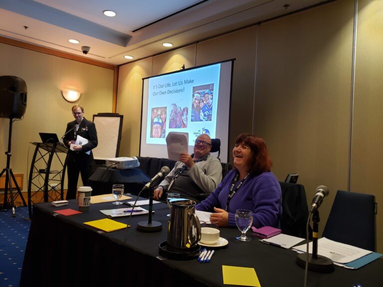 three people seated in a room at a table with a projector screen behind them. There are microphones on the table and papers in front of the people. They are smiling and laughing.