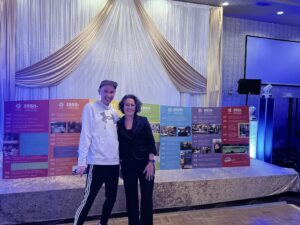 A man and a woman stand in front of a stage decorated with gold and white sashes. large colourful standing posters sit on the stage. Each poster has a header noting a range of years and a list of accomplishments and activities of Mission Association for Community Living over the years. 