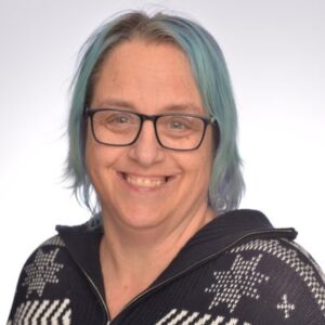 A portrait of a woman smiling at the camera. She has teal hair and black-rimmed glasses. She is wearing a black zip up sweater with white patterns.