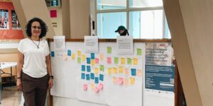 A woman wearing a white T-shirt stands in a large room beside a white board with many colourful sticky notes on it. The notes have writing on them and are under pieces of paper with writing prompts.