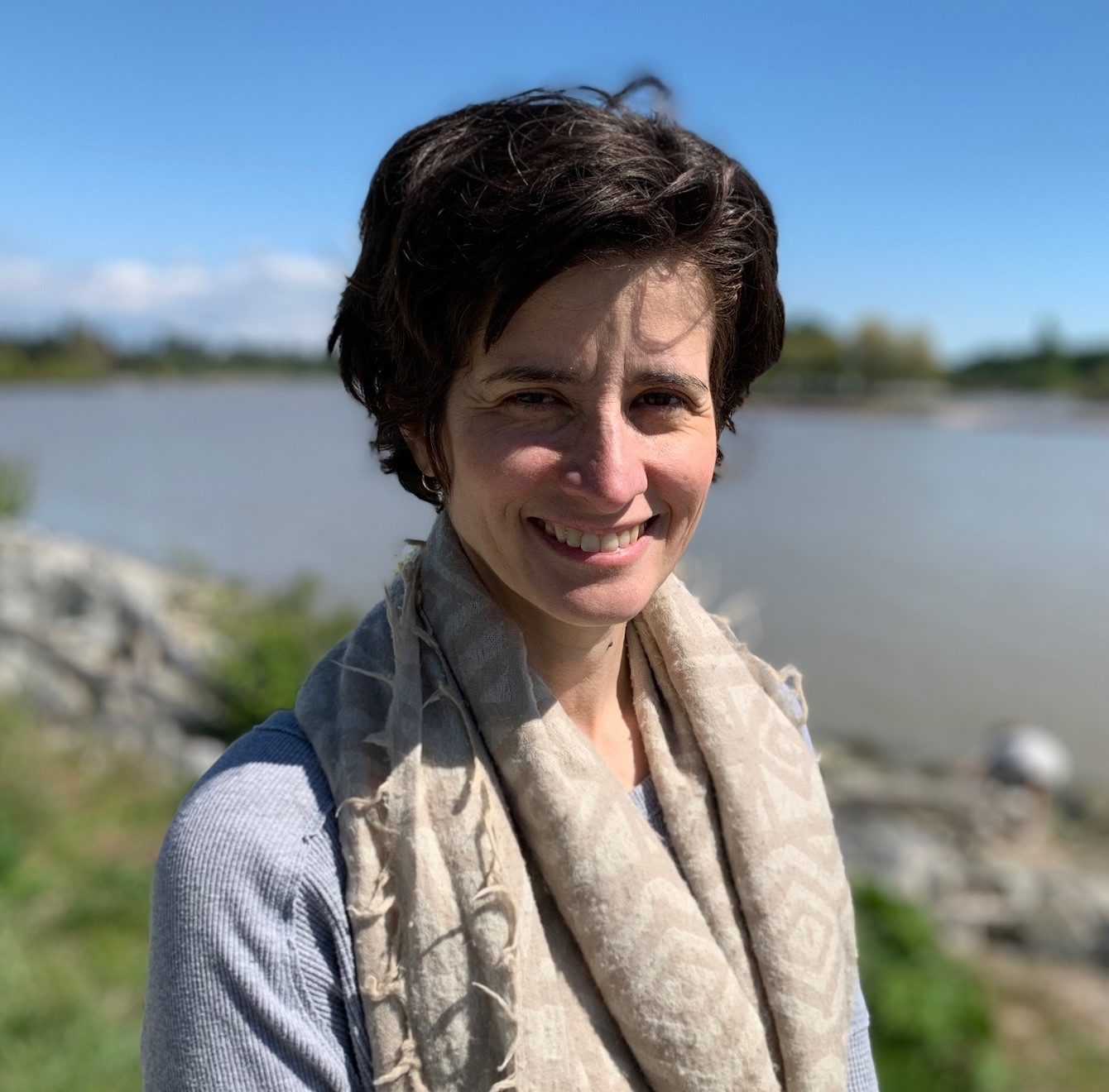 Hilary is smiling and has short dark brown wavy hair. She is wearing a grey long sleeved shirt with a beige scarf that has a cream colored diamond pattern draped around her neck. The background is somewhat blurry and has bright green grass and grey rocks next to a grey-blue river with a light blue sky.