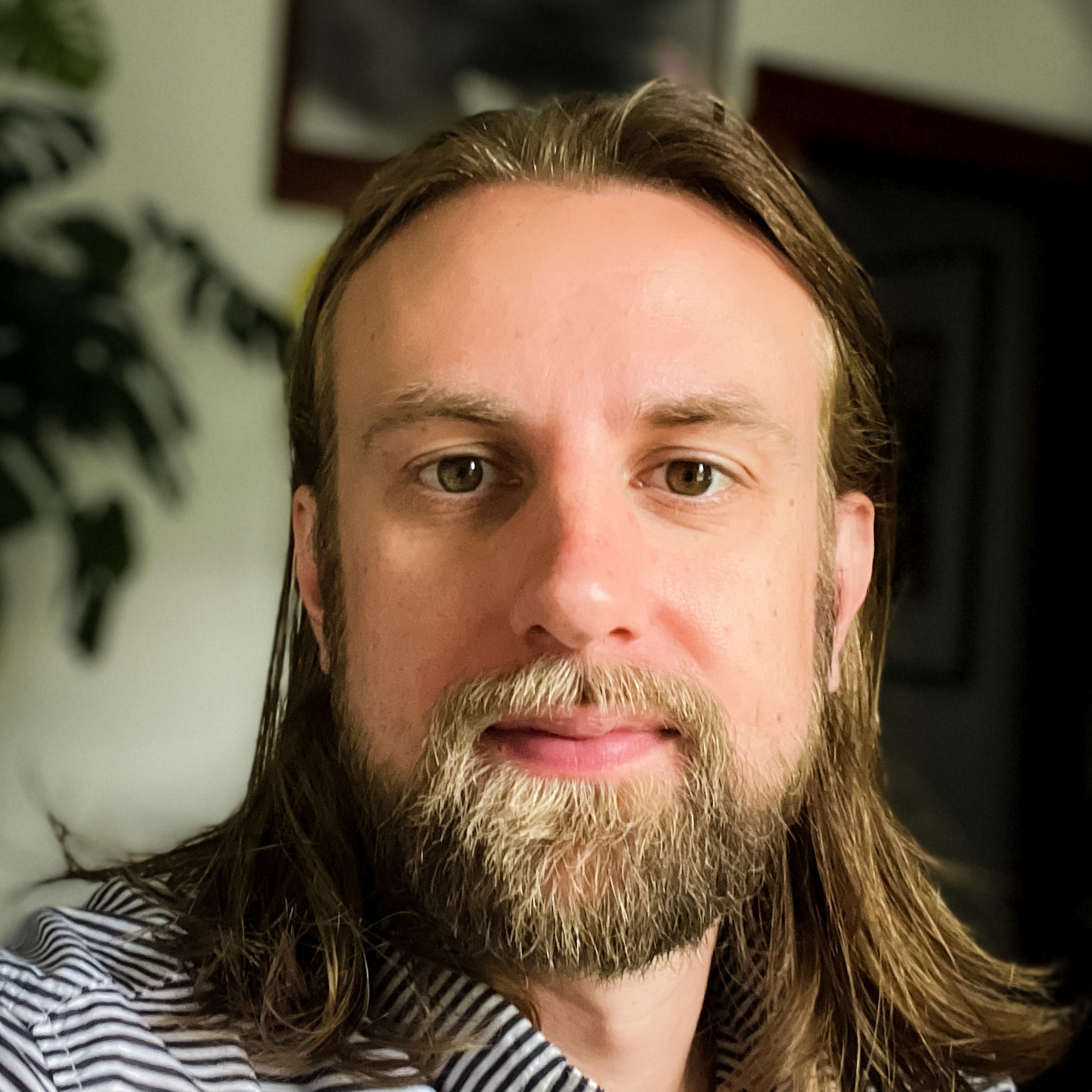 a man with shoulder length brown hair and styled facial hair. He is wearing a black and white striped button-up shirt and is in a room with low lighting and a monstera plant visible in the background