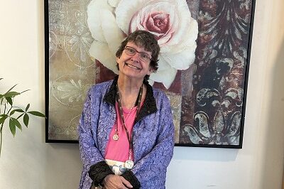 An elderly woman with short brown hair stands in front of a large painting of a white rose. She is wearing glasses, and a purple jacket over a pink shirt