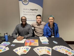 Three men at an exhibit table with pamphlets and info sheets spread across a black table. A banner is in the background that reads: poverty reduction coalition. working together for a poverty free british columbia