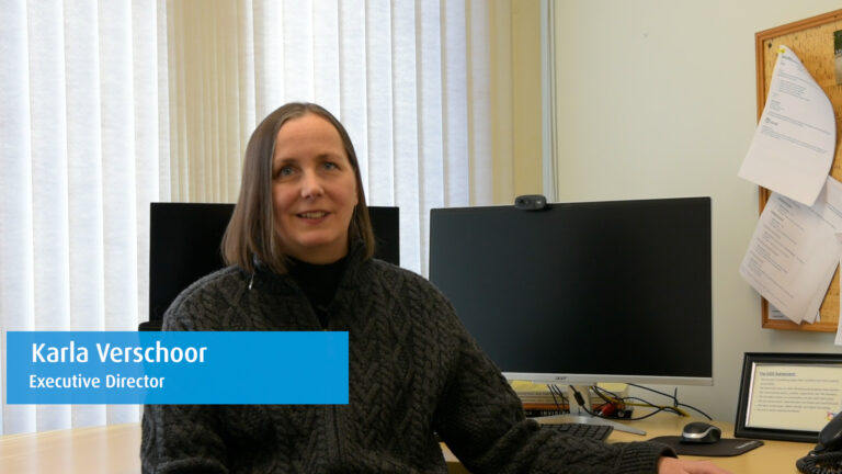 A woman with long brown hair seated in an office. She is smiling and speaking. There is a label to the bottom left of the screen that reads: Karla Verschoor. Executive Director.
