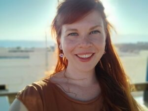 Kelsey Savage. a woman with long red hair who is outside on a sunny day wearing an orang short-sleeved shirt