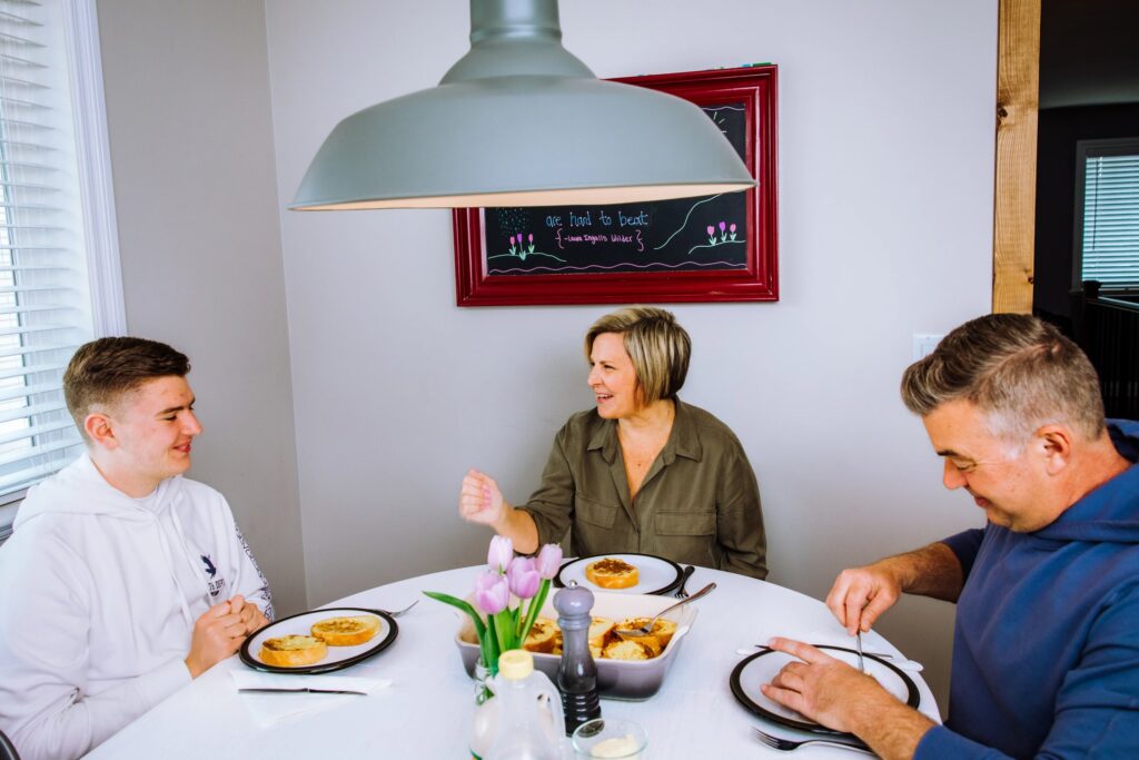 Two parents and their teenage son sit at a white dining table with dinner in front of them. There is a small arrangement of flowers in the centre of the table.