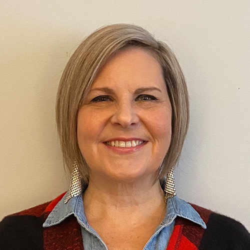 a woman with shoulder length blonde hair and gold earrings. she is wearing a denim button up shirt under a red plaid top
