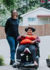 Two women outside on a neighborhood sidewalk. The person on the left has long brown hair, black glasses, a black sweater, and light blue jeans. The person on the right has short brown hair and is seated in a powered wheelchair and is wearing a burgundy zip up sweater over an orange top with black yoga pants. 