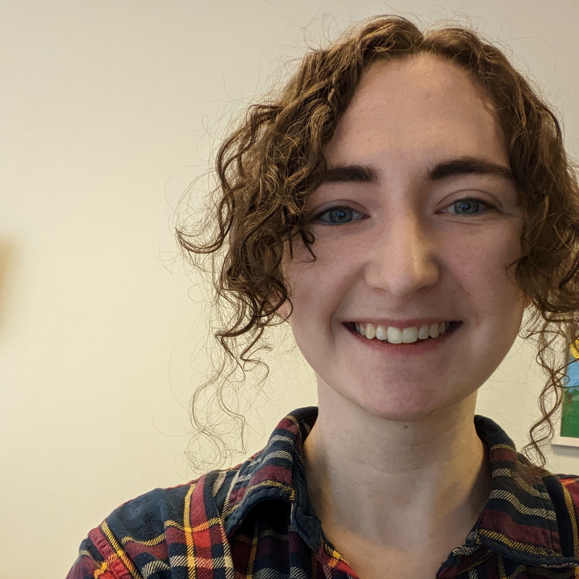 a young woman wearing a plaid shirt smiling at the camera. She has long curly brown hair tied up behind her head.