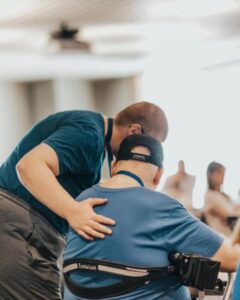 two people as seen from behind. One person has their hand on the back of the other person who is seated in a chair. They are both wearing blue shirts.
