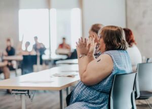 A woman with short brown hair wearing a blue dress is seated at a table at the front of a room of people. She has her hands up in front of her and is speaking.