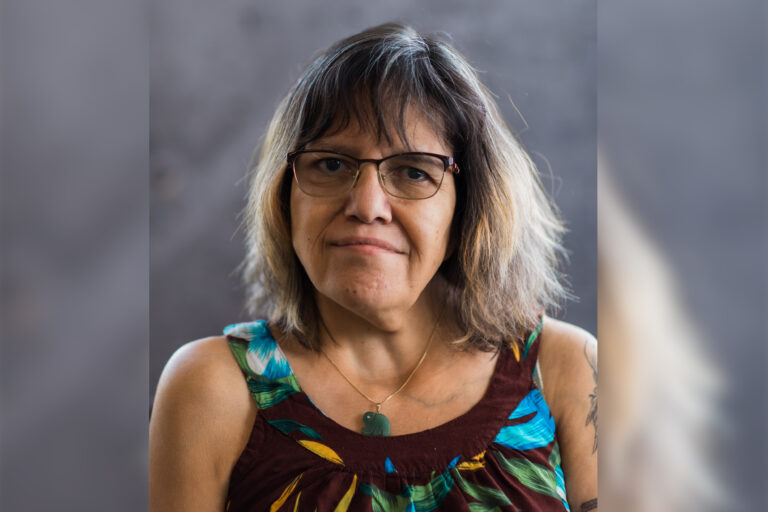 A portrait of a woman with shoulder length dark brown hair that is dyed a light straw colour closer to the ends. She is searing brown a gold rimmed glasses, a gold necklace with a green stone pendant and a brown floral sleeveless top.