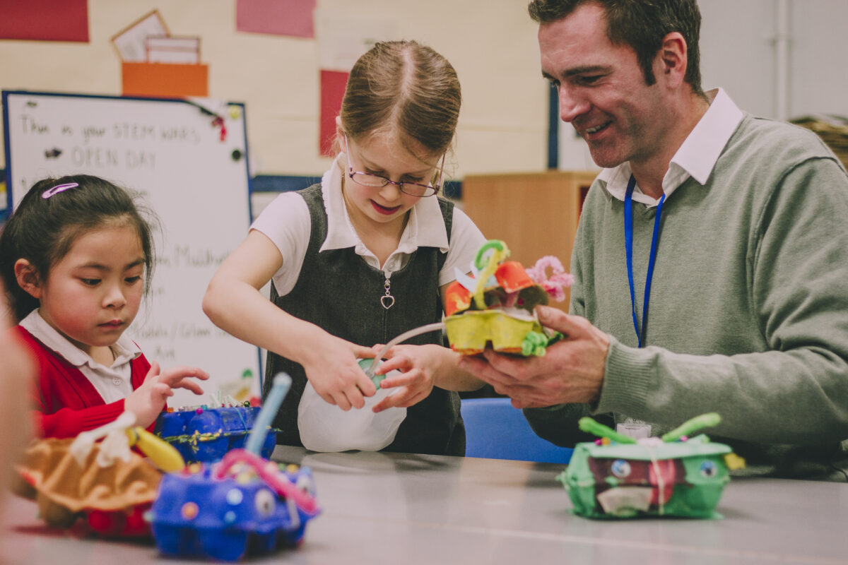 A young girl with blonde hair, a young girl with black hair, and a man with brown hair are in a classroom at a desk. The blonde child is holding a bottle with a tube that is attached to a painted and decorated egg carton that the man is holding. the child with black hair is looking at another decorated egg carton in front of her.