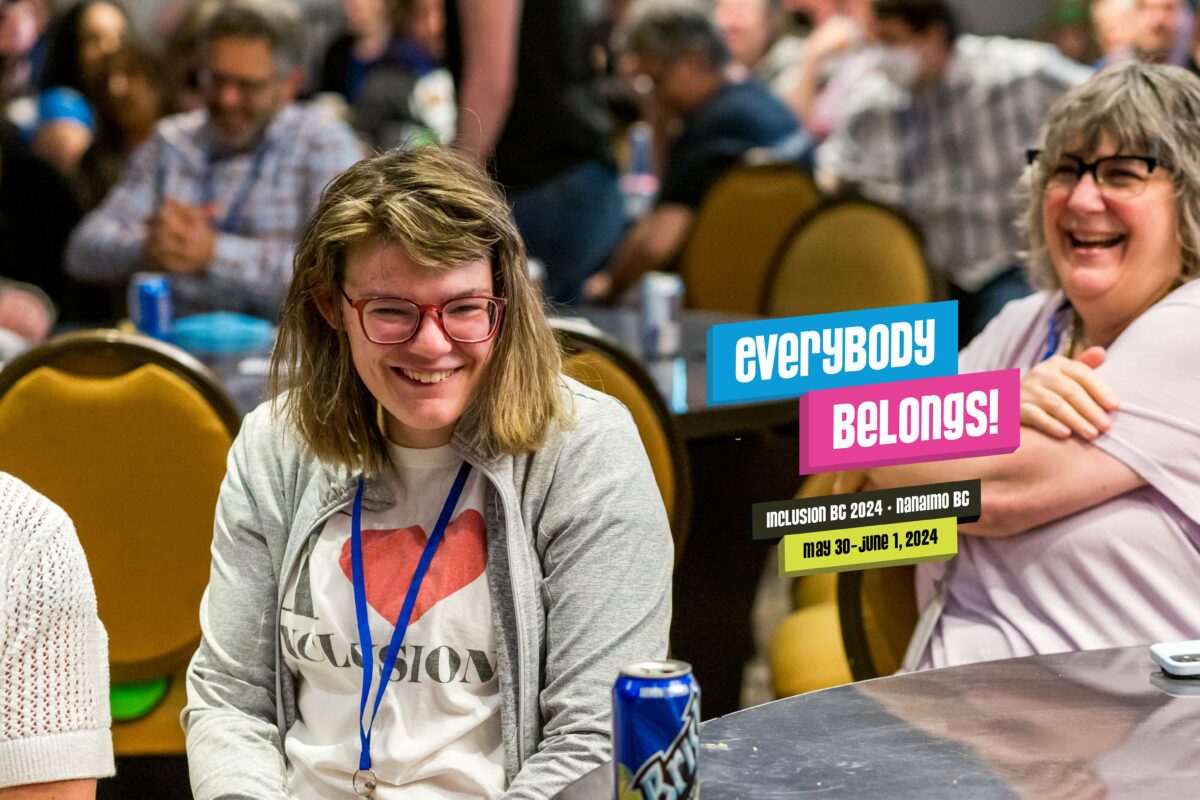 A young woman with long blonde hair and red glasses seated in a ballroom full of people at a large round table. She's wearing a white shirt that reads: I (heart symbol) Inclusion
