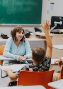 A young woman with shoulder length brown hair wearing a light blue button up sweater sits at the front of a classroom facing students seated at desks. One of the students is holding their hand up in the air. 