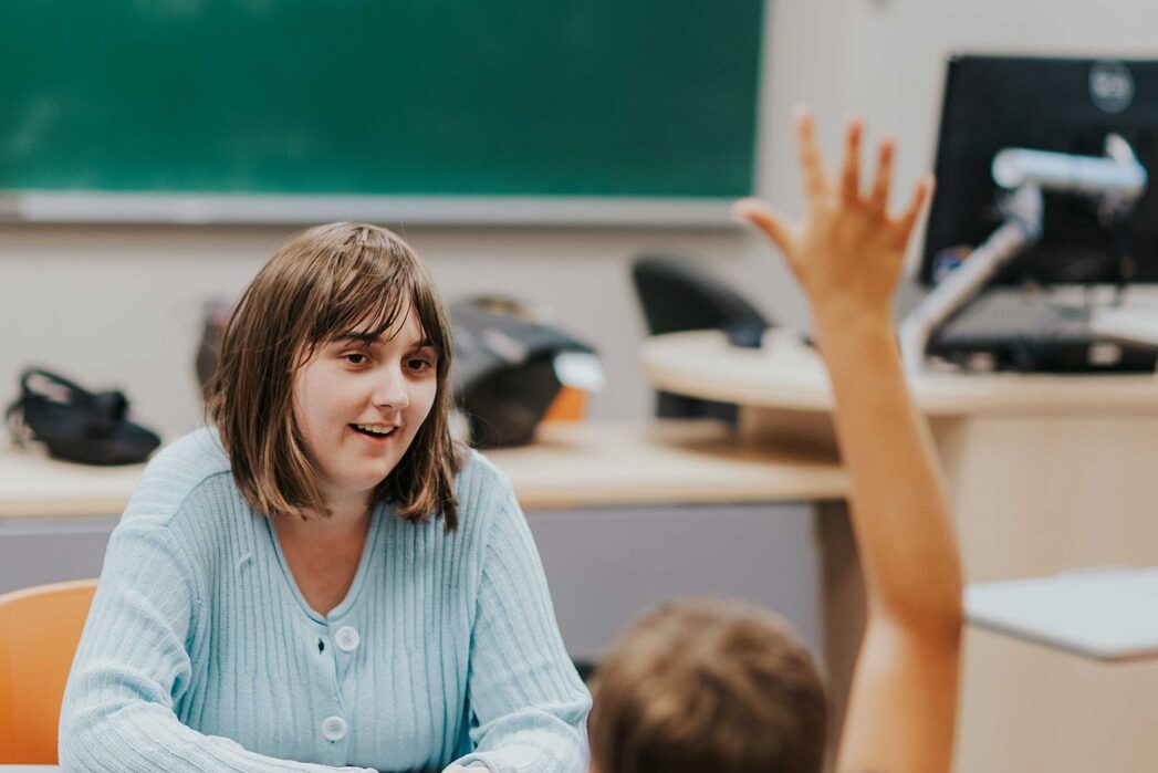 A young woman with shoulder length brown hair wearing a light blue button up sweater sits at the front of a classroom facing students seated at desks. One of the students is holding their hand up in the air.