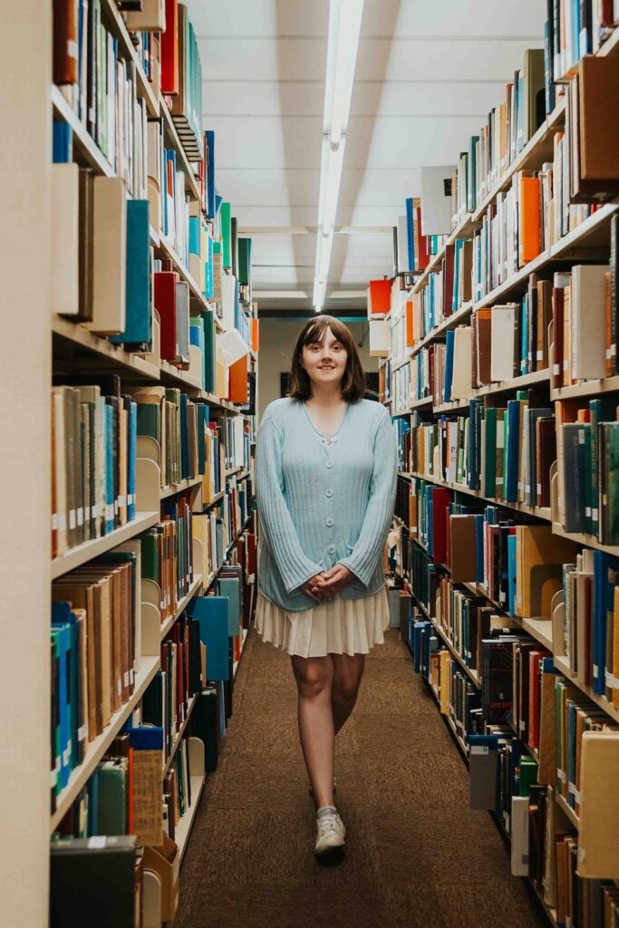 A young woman with shoulder length brown hair, a light blue button up sweater, and a white skirt stands between two library shelves. She is holding her hands together and has one foot behind the other mid-stride. 