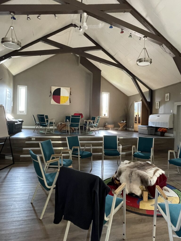 An indoor community gathering space with two circles of chairs around large drums covered with ornate blankets. The space has high ceilings and is low lit by bright sunlight shining through two tall windows on the back wall.