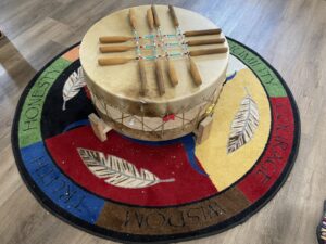 A large animal skin drum with seven drum sticks laying in a cross pattern over the top. The drum is sitting on a round rug not much larger than the drum itself. The rug is colourful decorated with the images of feathers. The words honesty, truth, wisdom, courage, and humility wrap the outer edge of the circular rug