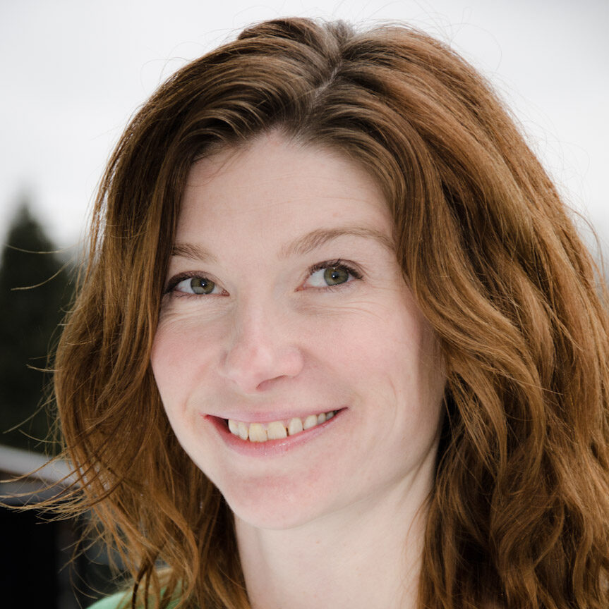 A woman with light skin, rosy cheeks, wavy long brown hair. She is outside on a cloudy day and is smiling at the camera. She is wearing a light green shirt.