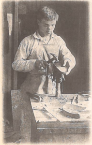 a black and white image on newspaper. A young man wearing a white shirt and an apron stands at a workshop table working on a shoe with a hammer in one hand.