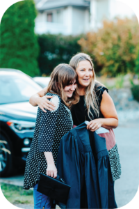 a young woman leans into a woman a little older than her while being hugged from the side. The older woman is smiling and looking into the distance and holding a graduation robe in one hand for the young woman. The young woman is holding her graduation cap in one hand. They both have light blonde/brown hair. 