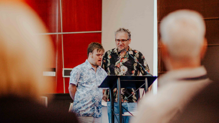 Two men stand at a podium in front of people seated in a large hotel conference room.