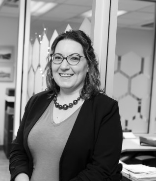 A black and white image of Heather who is smiling. She has shoulder length wavy hair, is wearing a plain colored top and a dark blazer, a beaded necklace, and plastic rimmed glasses. The background is an office with glass panel walls. The glass has frosted hexagon shapes on it and there is a desk in the office.