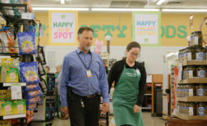 Two employees walking through a grocery store. The man on the left is wearing a light blue button up shirt and the woman on the right is wearing a green apron over a black long sleeved shirt.