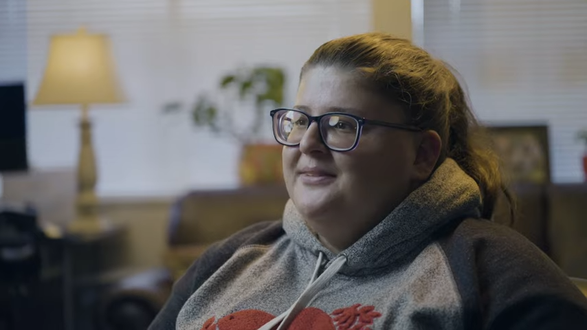 A young woman with glasses and brown hair in a ponytail looking to the side. She is wearing a two tone grey hoodie with a red logo on the front