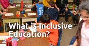 An image of a classroom full of children at their desks writing on paper. Some are smiling and working together. There is white text over the image that reads: What is Inclusive Education?