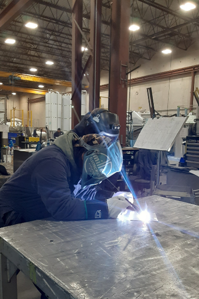 A person in a large industrial workshop wearing a welding mask and welding on the top of a large metal workbench. The welding is shining a bright light over the person's mask and body. 
