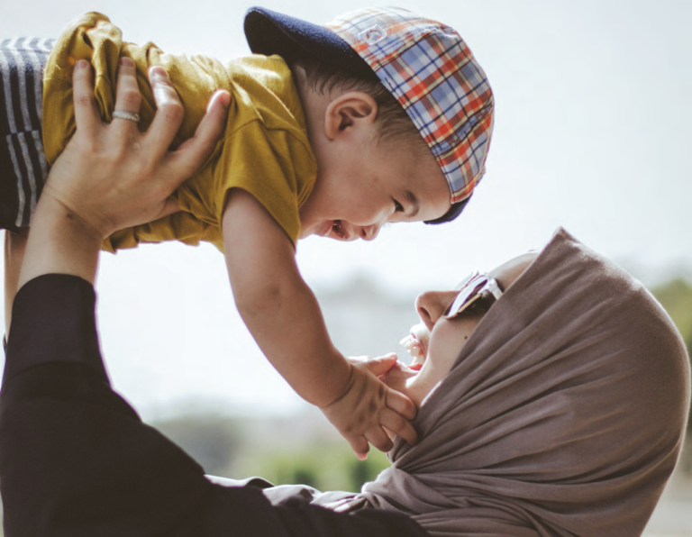 A woman wearing a head covering and sunglasses and her small child who is wearing a yellow t shirt and backwards baseball cap. They are smiling at each other while she holds the child up in the air.