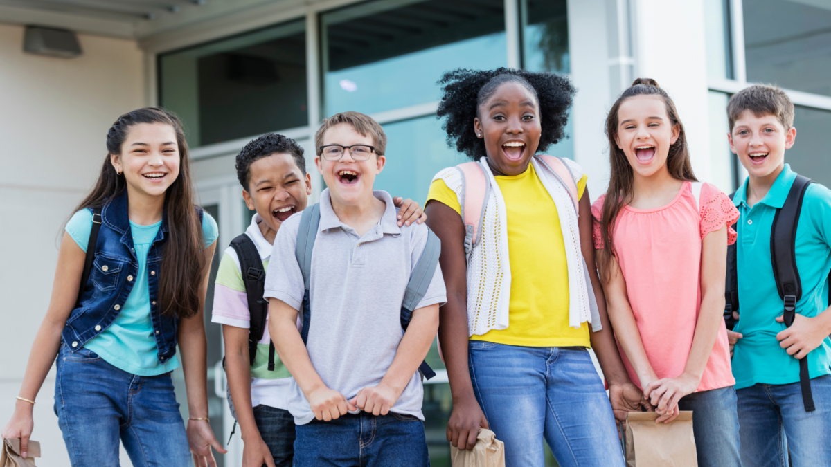 A diverse group of children in front of a school wearing backpacks.