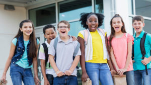 A diverse group of children in front of a school wearing backpacks.